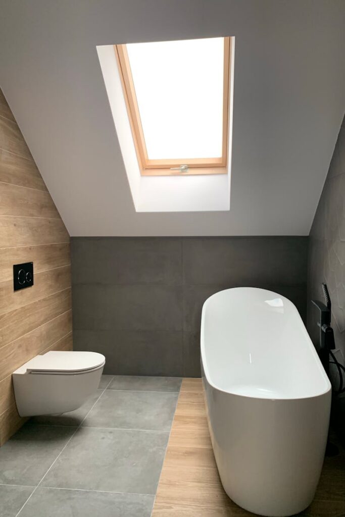 Modern interior of stylish minimal attic bathroom with white bathtub and toilet and roof window